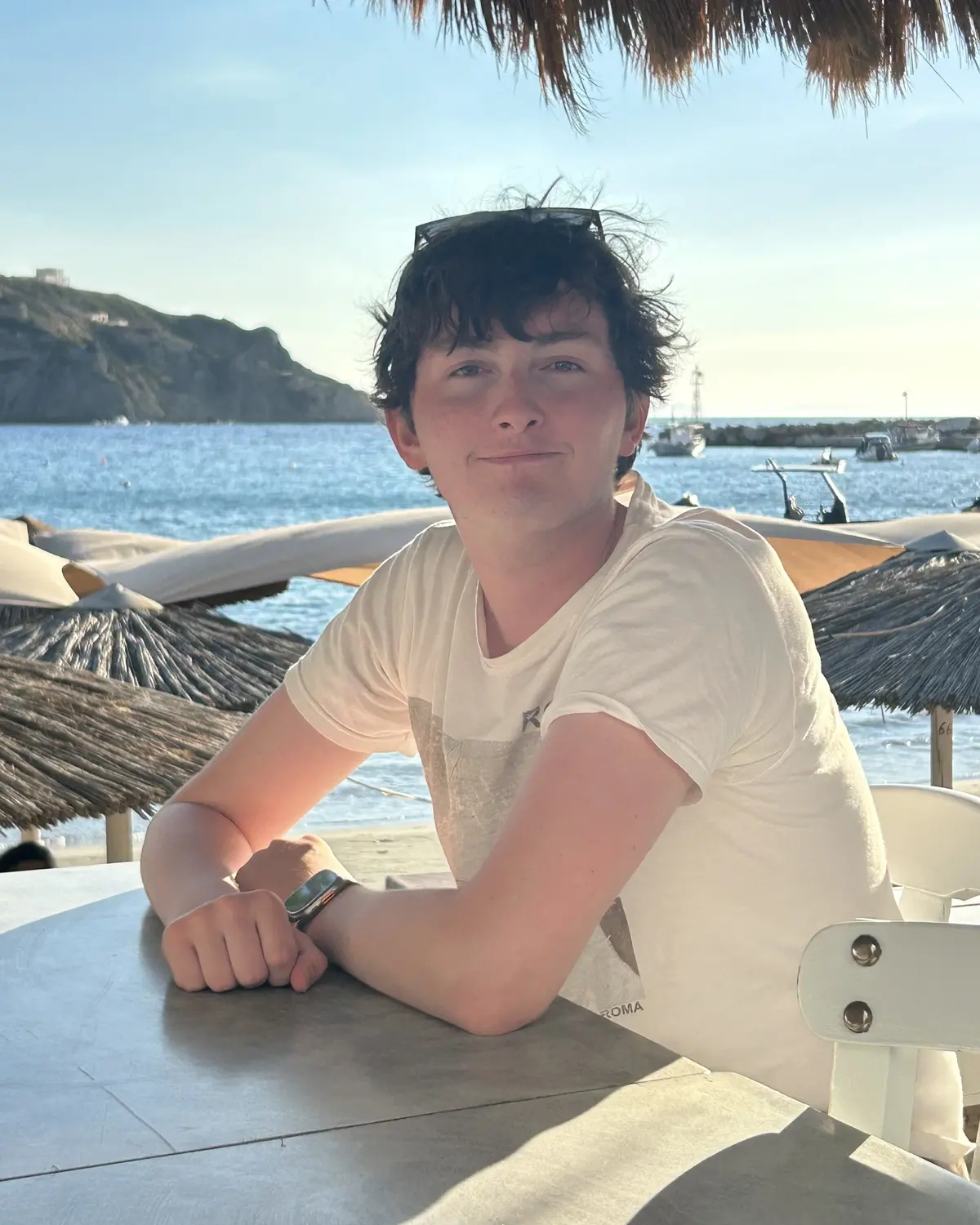 Alex Szeremeta at a seaside café table, wearing a white t-shirt and smiling.